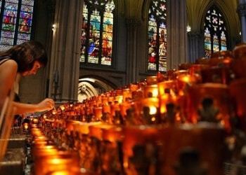 A dark-haired petitioner lights a votive candle in a cathedral, with stained-glass windows visible in the background behind a long row of lit votives.